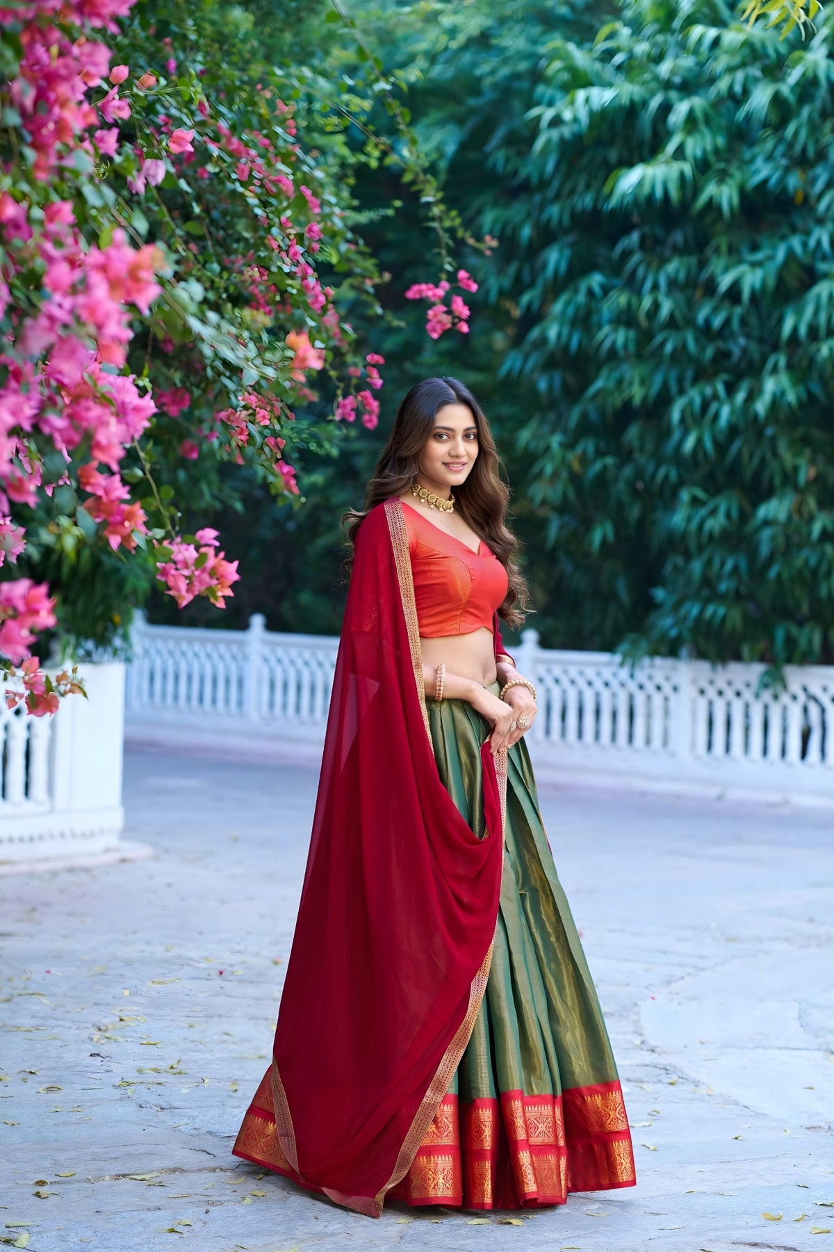 Woman in a red and green traditional outfit standing outdoors with pink flowers and greenery.
