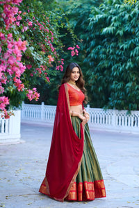 Woman in a red and green traditional outfit standing outdoors with pink flowers and greenery.