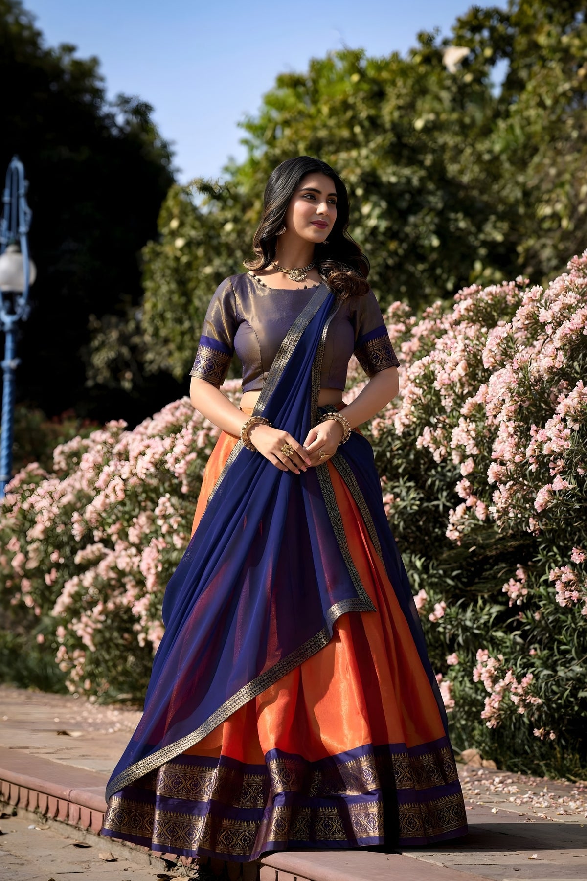 Woman in a traditional saree standing outdoors with flowers and trees in the background