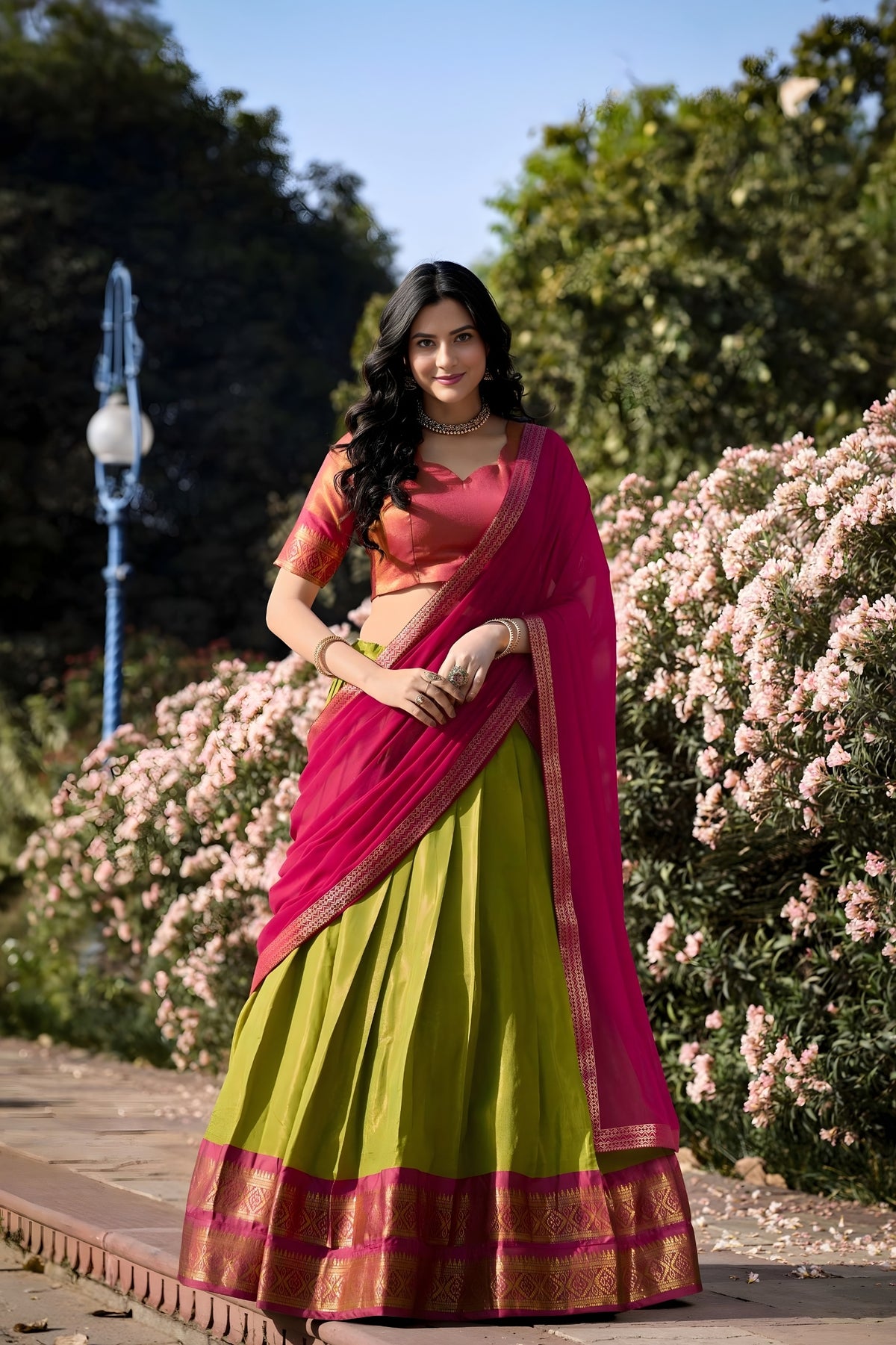 Woman in a green and pink saree standing in a garden with flowers and trees.