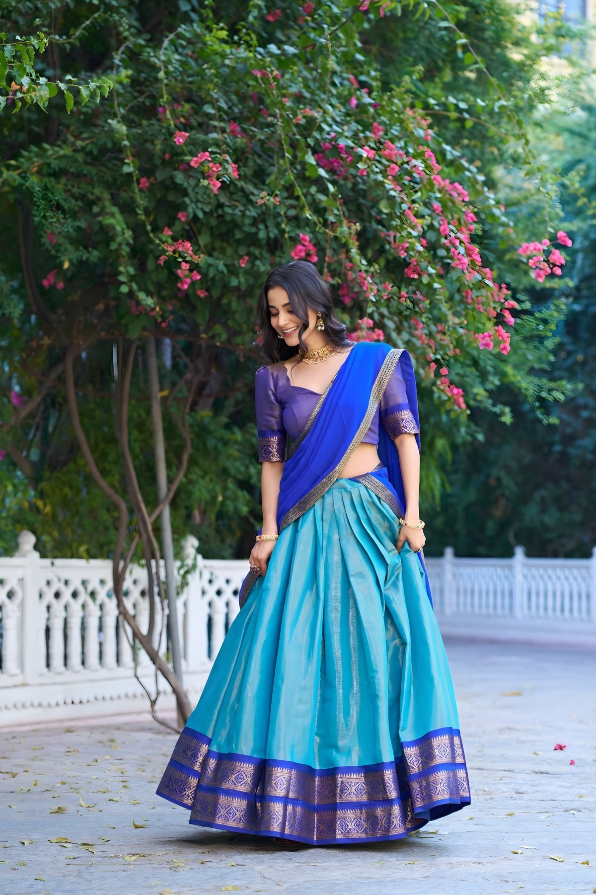 Woman in a blue and purple traditional outfit standing in a garden with greenery and flowers.