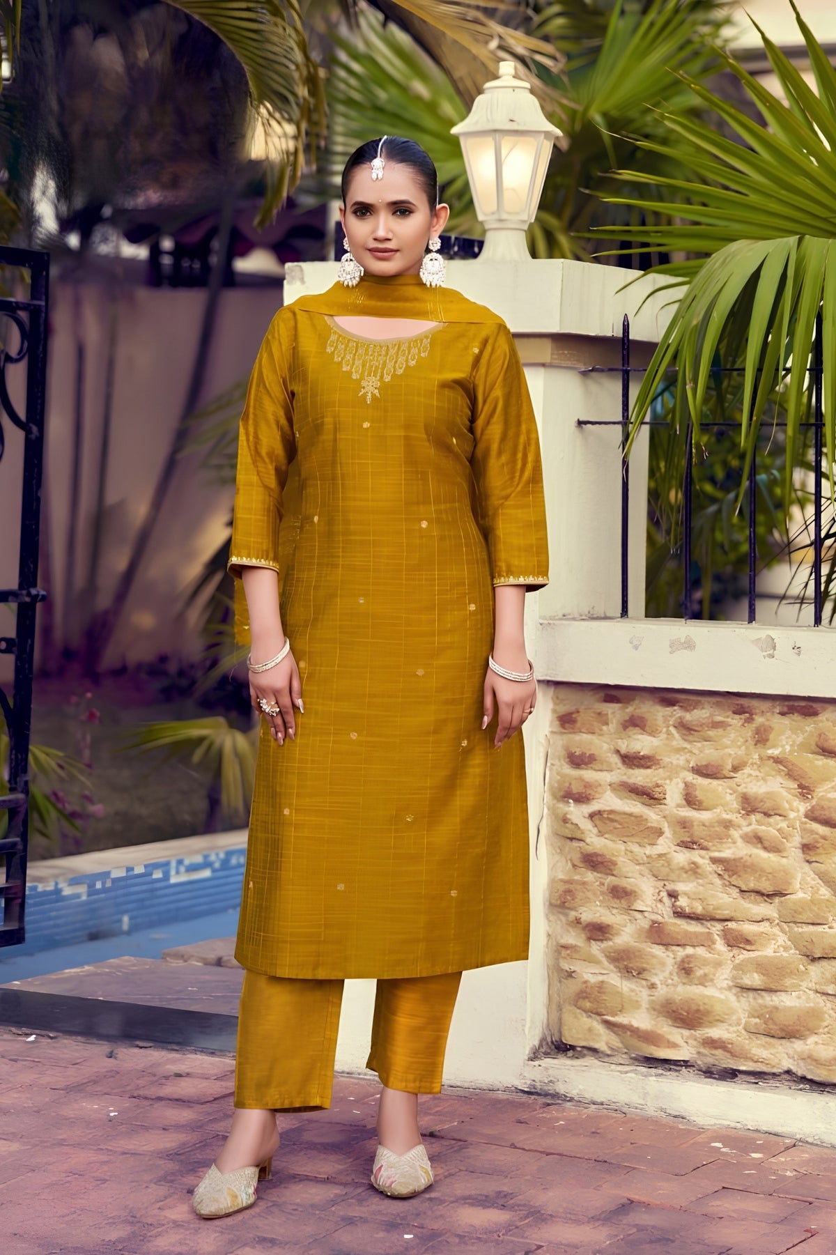 Woman wearing a mustard yellow traditional outfit standing outdoors with plants and a stone wall in the background.