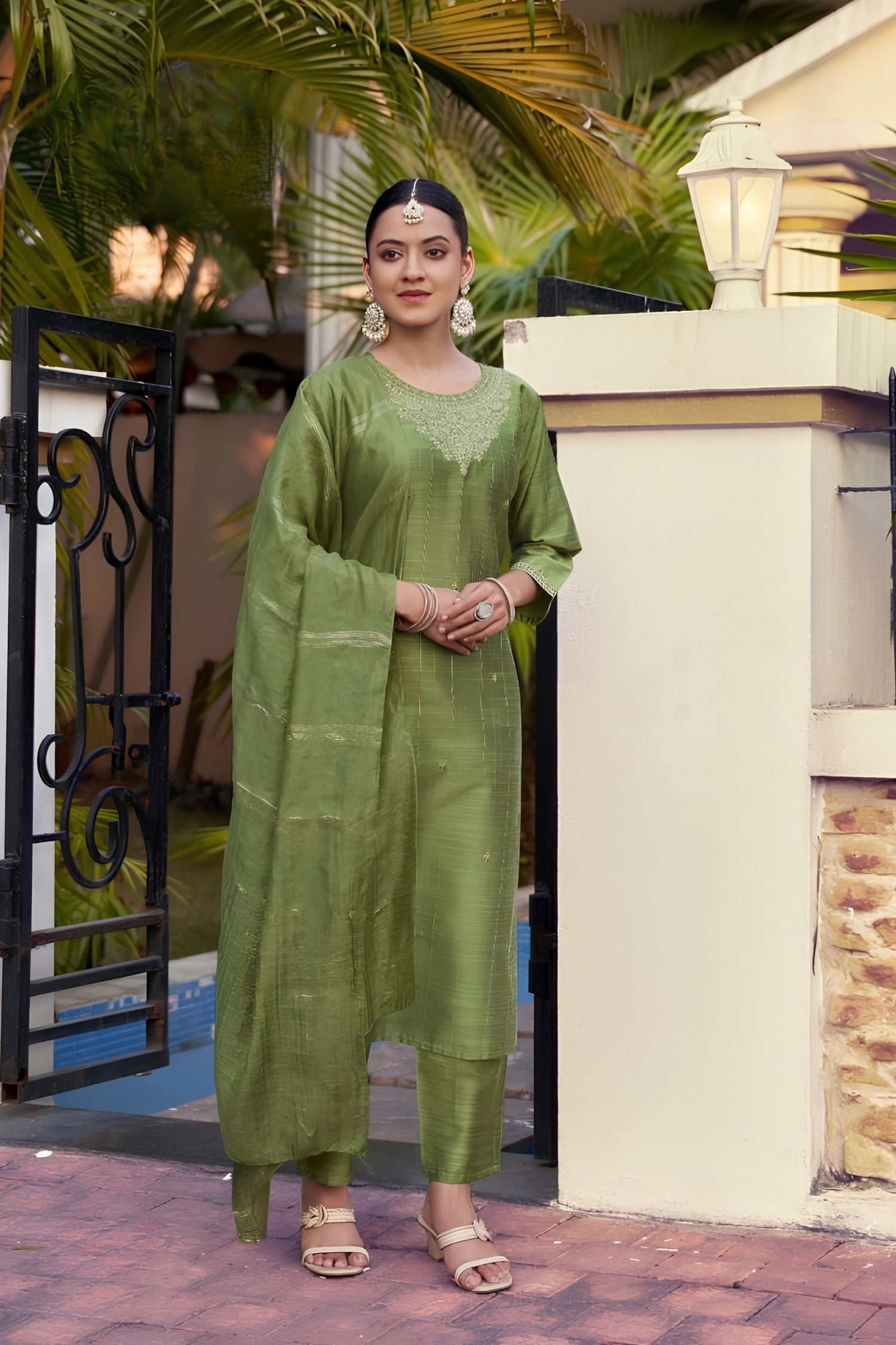 Woman in a green traditional outfit standing outdoors with plants and a building in the background.