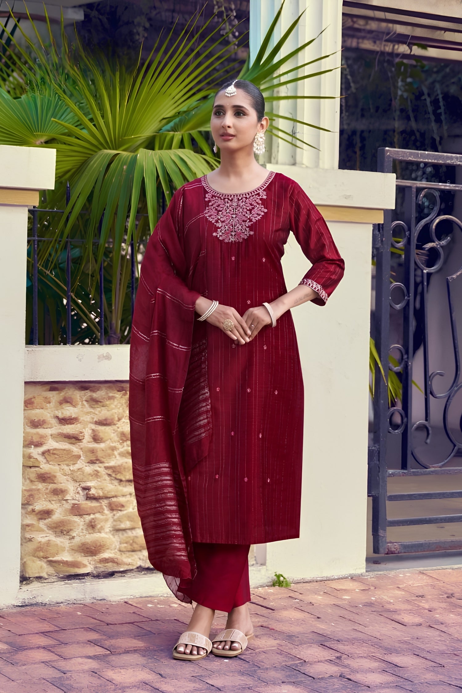 Woman in a red traditional outfit standing outdoors with greenery and a building in the background.