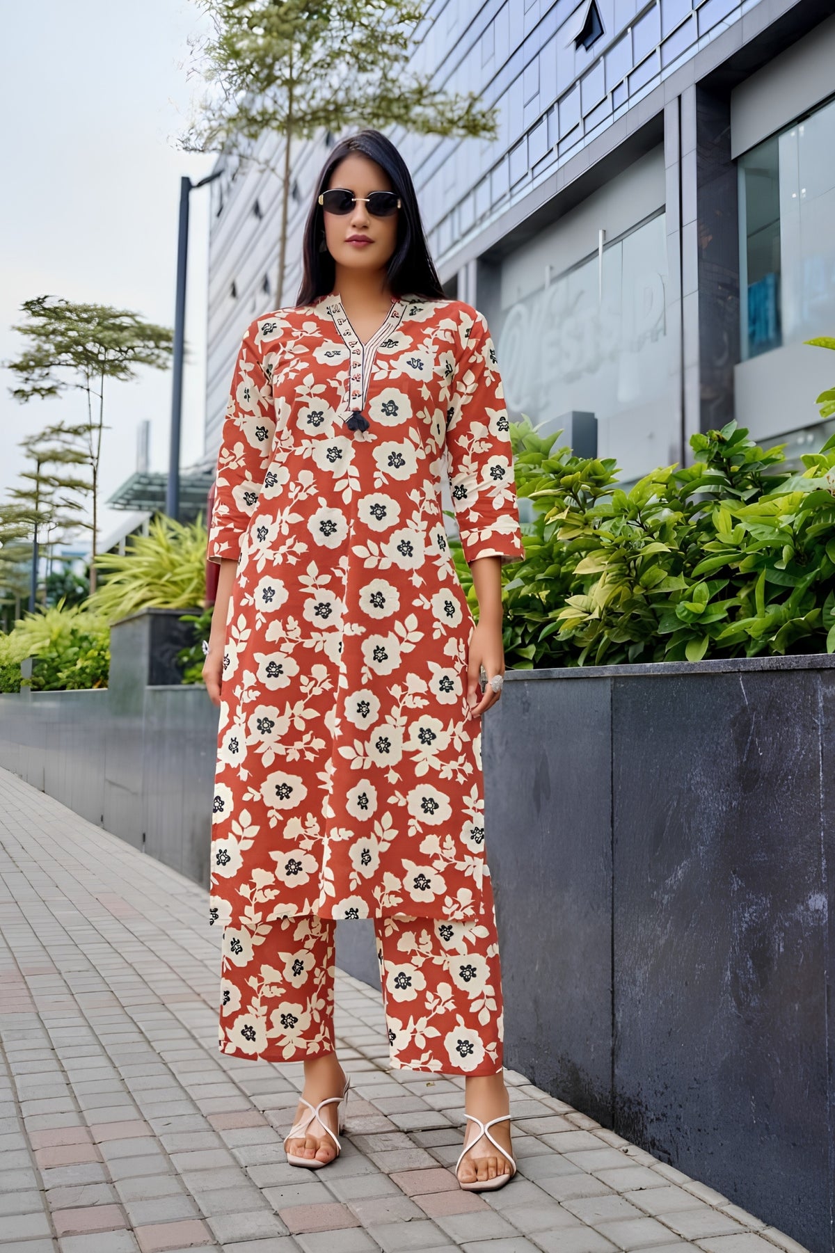 Woman wearing a red floral jumpsuit standing outdoors with greenery and a building in the background.