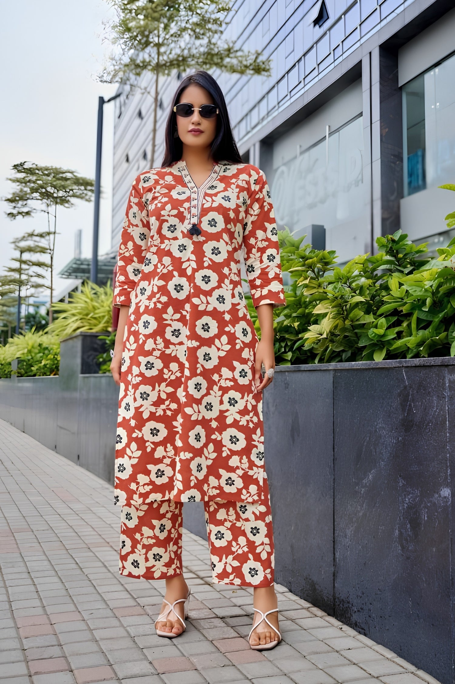 Woman wearing a red floral jumpsuit standing outdoors with greenery and a building in the background.